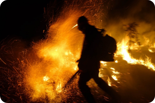 Brigadista em silhueta no meio de um incêndio florestal à noite. As chamas e fagulhas intensas dominam a cena, criando um contraste dramático com o corpo escuro do profissional que atua com ferramenta em mãos em meio ao fogo.