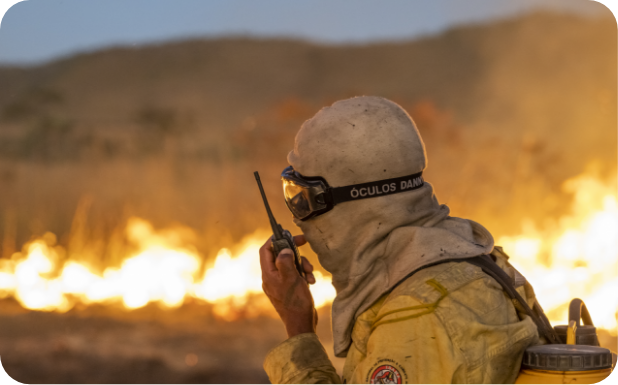 Brigadista com uniforme de proteção amarela e máscara comunica-se por rádio durante o combate a um incêndio florestal. O fogo consome a vegetação ao fundo, iluminando a cena ao entardecer. A faixa no capacete traz a inscrição “ÓCULOS DAMA”.