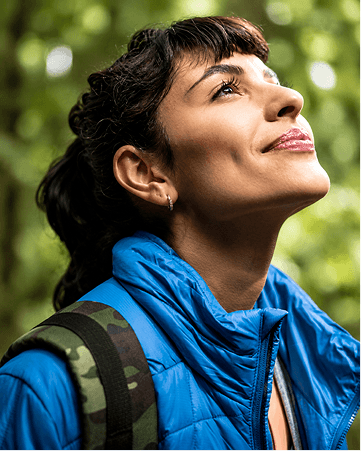 Uma mulher sorridente com traços latinos é retratada do busto para cima, olhando com admiração em direção ao céu. Ela tem cabelos escuros e ondulados presos em um rabo de cavalo alto com uma franja reta sobre a testa, usa um pequeno brinco de argola dourada e exibe uma expressão serena com lábios pintados por um gloss. Sua vestimenta é composta por uma jaqueta de nylon acolchoada em azul-bic vibrante sobreposta a uma regata verde-clara, e traz no ombro direito a alça larga de uma mochila com estampa de camuflagem militar em tons de verde, marrom e preto.